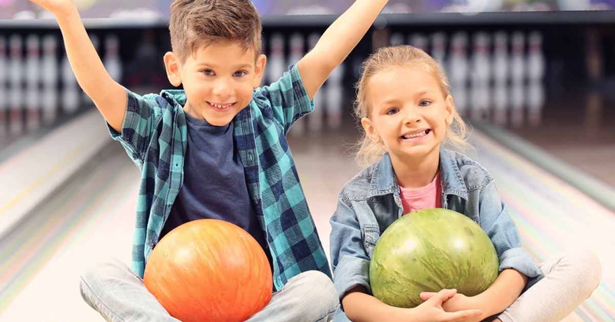 two young kids holding bowling balls, sitting on the lanes